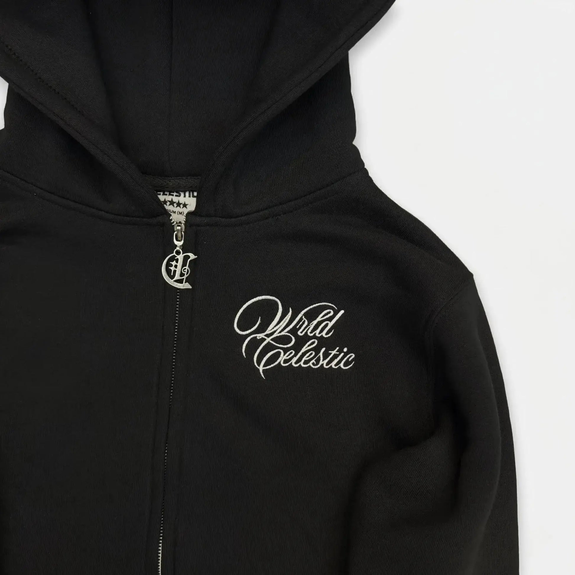Detailed close-up of the CELESTIC Black Basic zip-up hoodie. Highlighting the premium heavyweight black cotton fleece material, the elegant white script logo embroidery on the chest, and the custom silver metal 'C' zipper pull.