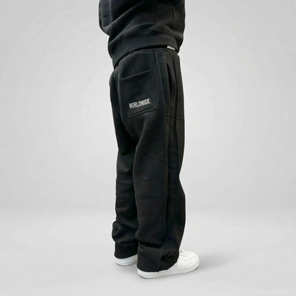 Back angle view of a model wearing the CELESTIC Black Basic sweatpants. These premium heavyweight black cotton fleece pants feature a relaxed straight-leg silhouette, prominent white 'WORLDWIDE.' embroidery on the right back pocket, and distinct side panel seams. Shown with white sneakers on a clean light background.