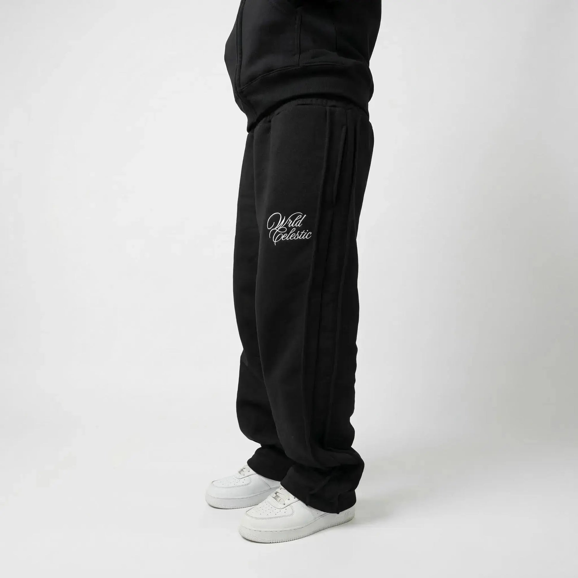 Side view of a model wearing the CELESTIC Black Basic sweatpants. These premium heavyweight black cotton fleece pants feature a relaxed straight-leg fit, prominent side panel seams, and elegant white script logo embroidery on the left thigh. Shown paired with white sneakers.