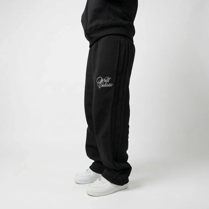 Side view of a model wearing the CELESTIC Black Basic sweatpants. These premium heavyweight black cotton fleece pants feature a relaxed straight-leg fit, prominent side panel seams, and elegant white script logo embroidery on the left thigh. Shown paired with white sneakers.