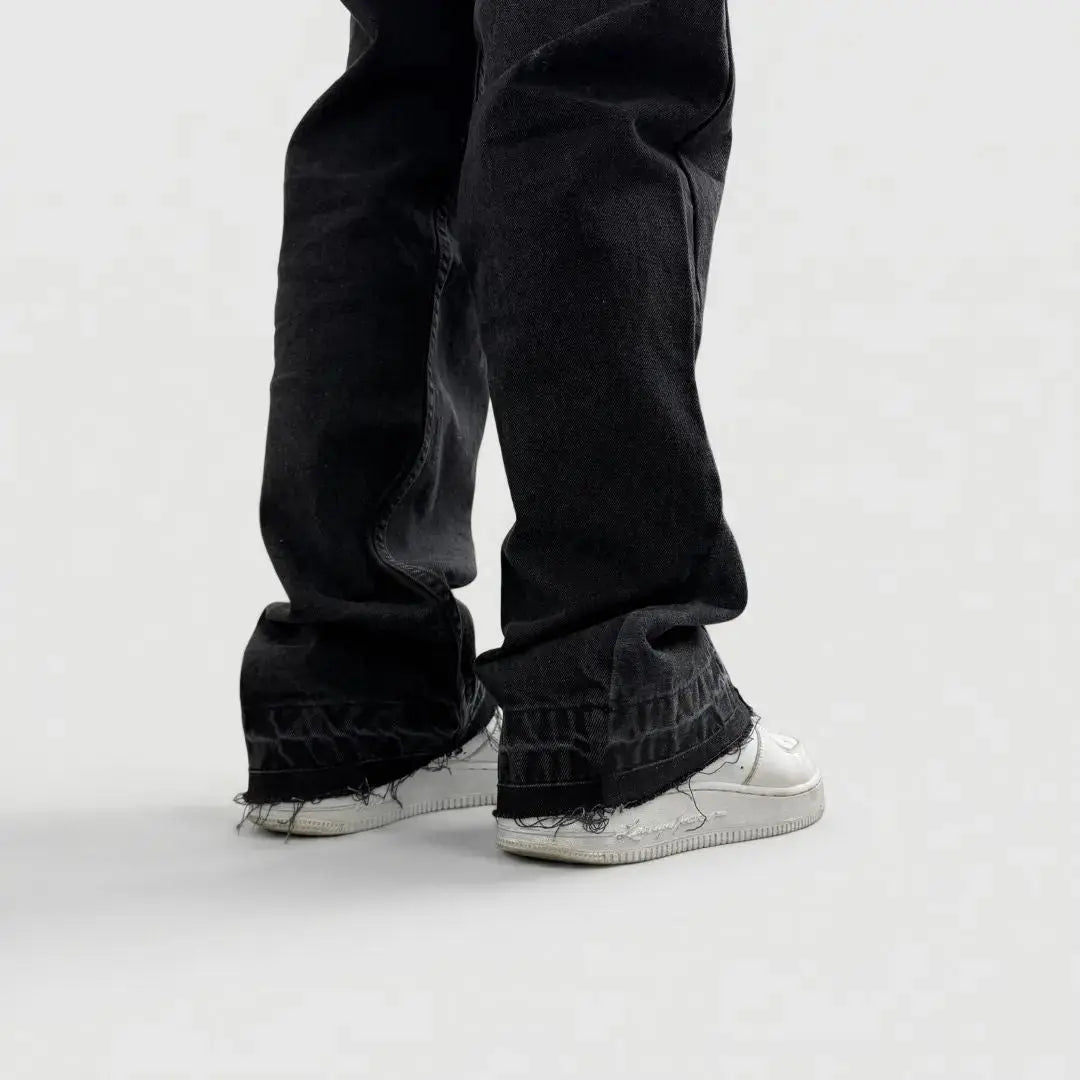 A detailed close-up rear view of the lower leg and hem of the CELESTIC Black Flared Denim Jeans on a white background. The image focuses on the intricate panelled construction of the flared leg and the heavily distressed, raw frayed edges at the bottom. The washed black denim texture is clearly visible as the fabric stacks over a white sneaker, showcasing the premium streetwear design.
