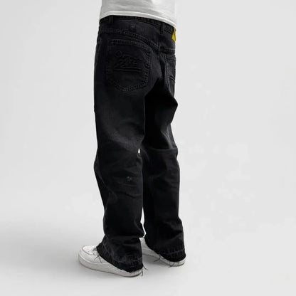 On-body rear view of the CELESTIC Black Flared Denim Jeans on a white background. The image showcases the detailed embroidery on the back pockets, the premium washed black denim fabric, and a yellow branded leather patch on the waistband. The relaxed flared silhouette and the raw, distressed hems at the bottom are clearly visible, styled with white sneakers.