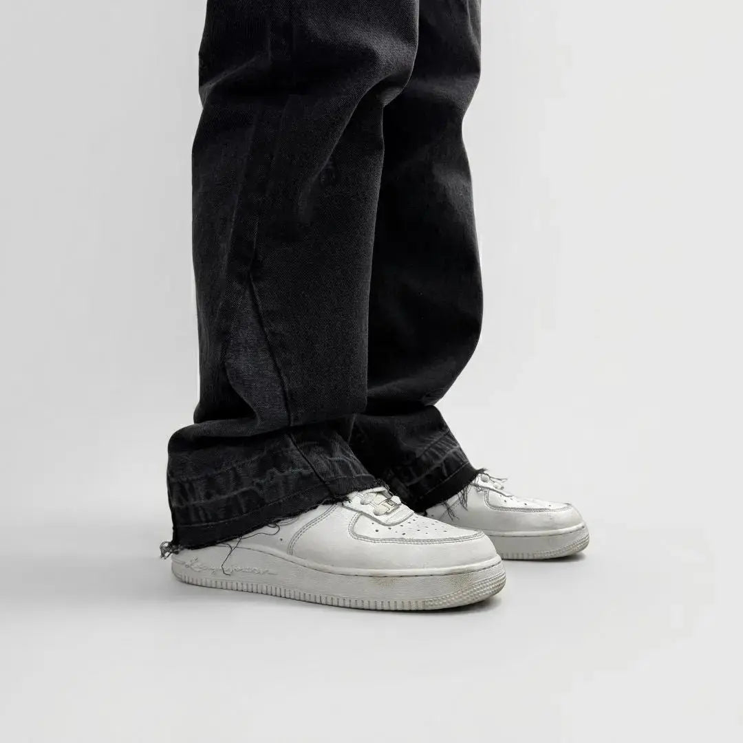 A detailed close-up photograph of the lower leg and hem of the CELESTIC Black Flared Denim Jeans, styled with white sneakers. The image highlights the premium washed black denim texture and the intricate flared-leg construction. The focus is on the raw, distressed, and frayed bottom hem, showcasing the rugged streetwear aesthetic and high-quality craftsmanship of the denim.