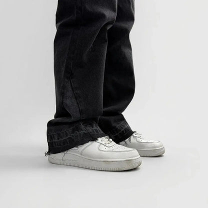 A detailed close-up photograph of the lower leg and hem of the CELESTIC Black Flared Denim Jeans, styled with white sneakers. The image highlights the premium washed black denim texture and the intricate flared-leg construction. The focus is on the raw, distressed, and frayed bottom hem, showcasing the rugged streetwear aesthetic and high-quality craftsmanship of the denim.