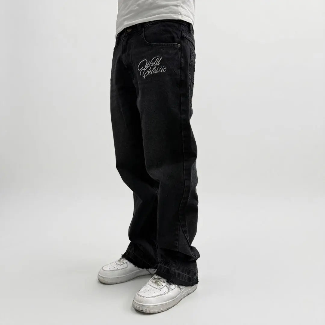 On-body side view of the CELESTIC Black Flared Denim Jeans styled with white sneakers. These premium black denim jeans feature a relaxed flared-leg fit with raw, distressed hems at the bottom. A white 'Wrld Celestic' script logo is embroidered on the left thigh. The photograph highlights the high-quality denim texture and the contemporary streetwear silhouette.