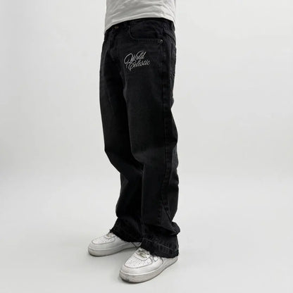 On-body side view of the CELESTIC Black Flared Denim Jeans styled with white sneakers. These premium black denim jeans feature a relaxed flared-leg fit with raw, distressed hems at the bottom. A white 'Wrld Celestic' script logo is embroidered on the left thigh. The photograph highlights the high-quality denim texture and the contemporary streetwear silhouette.