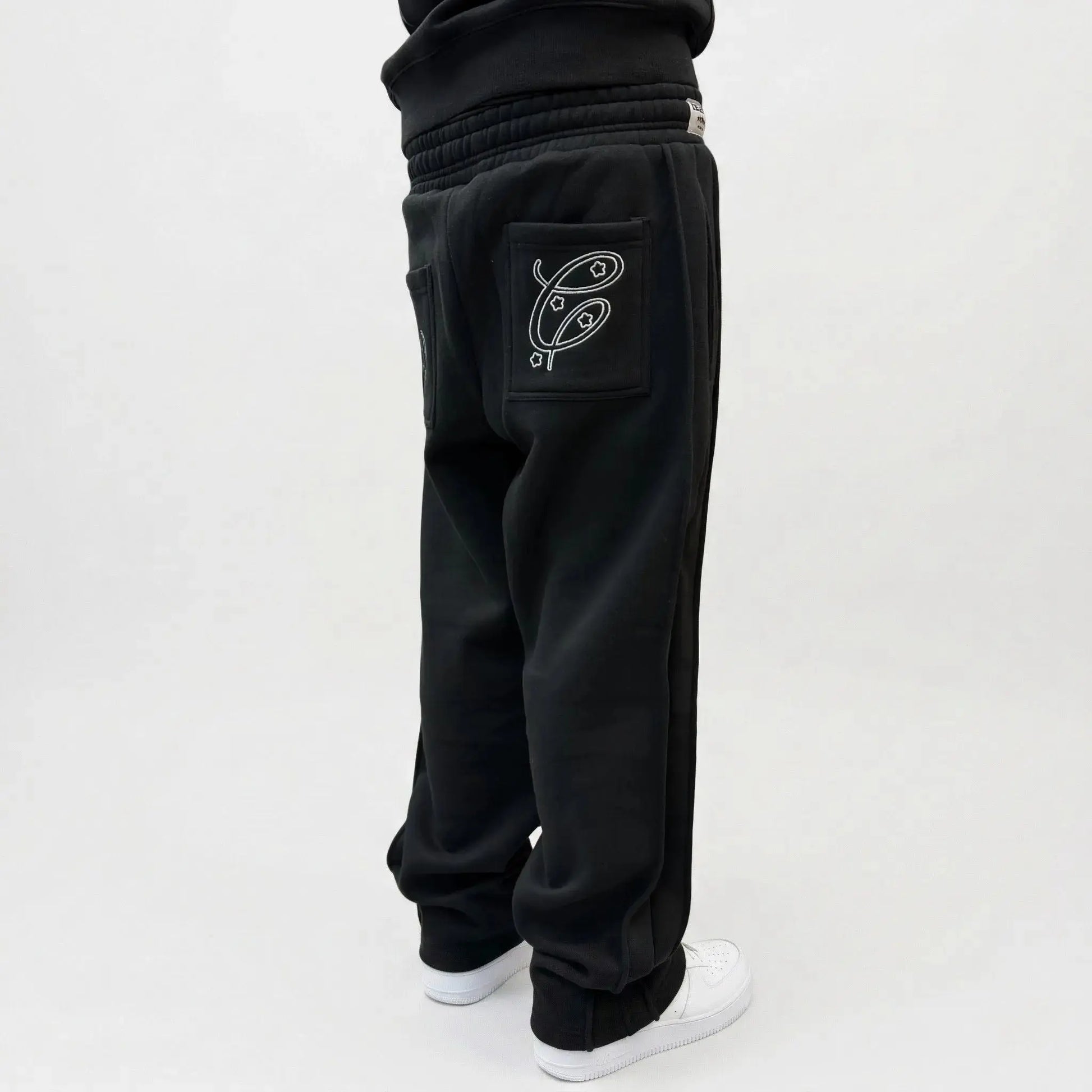 Black oversized tracksuit pants detailed close-up of cuffed ankle. Features the small white Celestic embroidered logo, ribbed cuff, white contrast stitching, and premium heavyweight fleece fabric texture, paired with black sneakers on a concrete background.