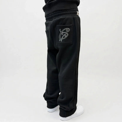 Black oversized tracksuit pants detailed close-up of cuffed ankle. Features the small white Celestic embroidered logo, ribbed cuff, white contrast stitching, and premium heavyweight fleece fabric texture, paired with black sneakers on a concrete background.