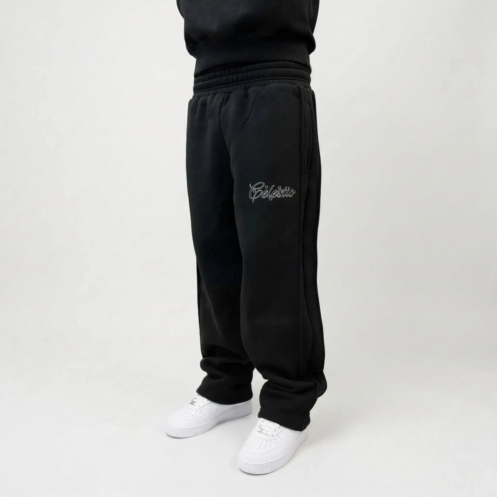 Black oversized tracksuit pants front angle view on model. Premium heavyweight cotton fleece sweatpants highlighting the white outline logo embroidery on the left thigh, side seam detailing, and the relaxed fit stacking perfectly over white sneakers.