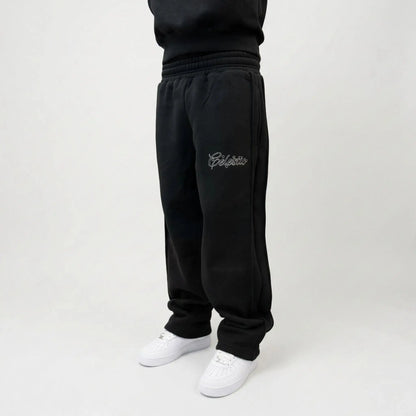 Black oversized tracksuit pants front angle view on model. Premium heavyweight cotton fleece sweatpants highlighting the white outline logo embroidery on the left thigh, side seam detailing, and the relaxed fit stacking perfectly over white sneakers.
