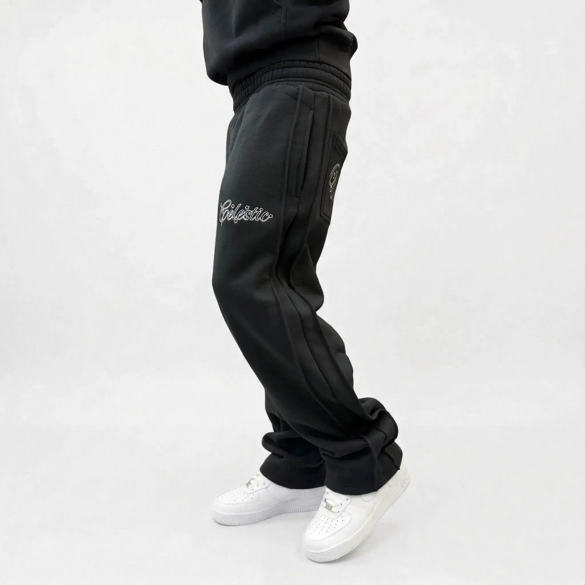 Black oversized tracksuit pants side view on model. Premium heavyweight cotton fleece sweatpants highlighting both the thigh and back pocket white outline logo embroidery, side seam detailing, and the relaxed fit stacking effortlessly over white sneakers.