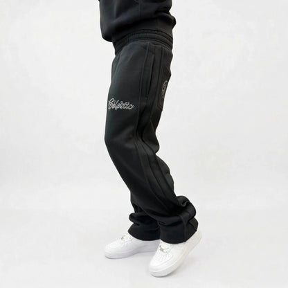Black oversized tracksuit pants side view on model. Premium heavyweight cotton fleece sweatpants highlighting both the thigh and back pocket white outline logo embroidery, side seam detailing, and the relaxed fit stacking effortlessly over white sneakers.