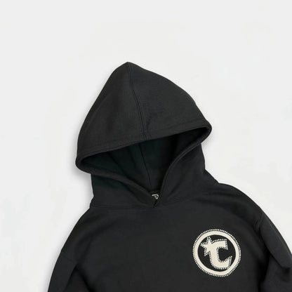 A close-up flat-lay photograph of the CELESTIC Black Star Hoodie. The image focuses on the upper section, highlighting the premium heavyweight black cotton fleece fabric and the structured double-lined hood. On the left chest, there is a prominent, circular white embroidered logo patch featuring a central five-pointed star and text.