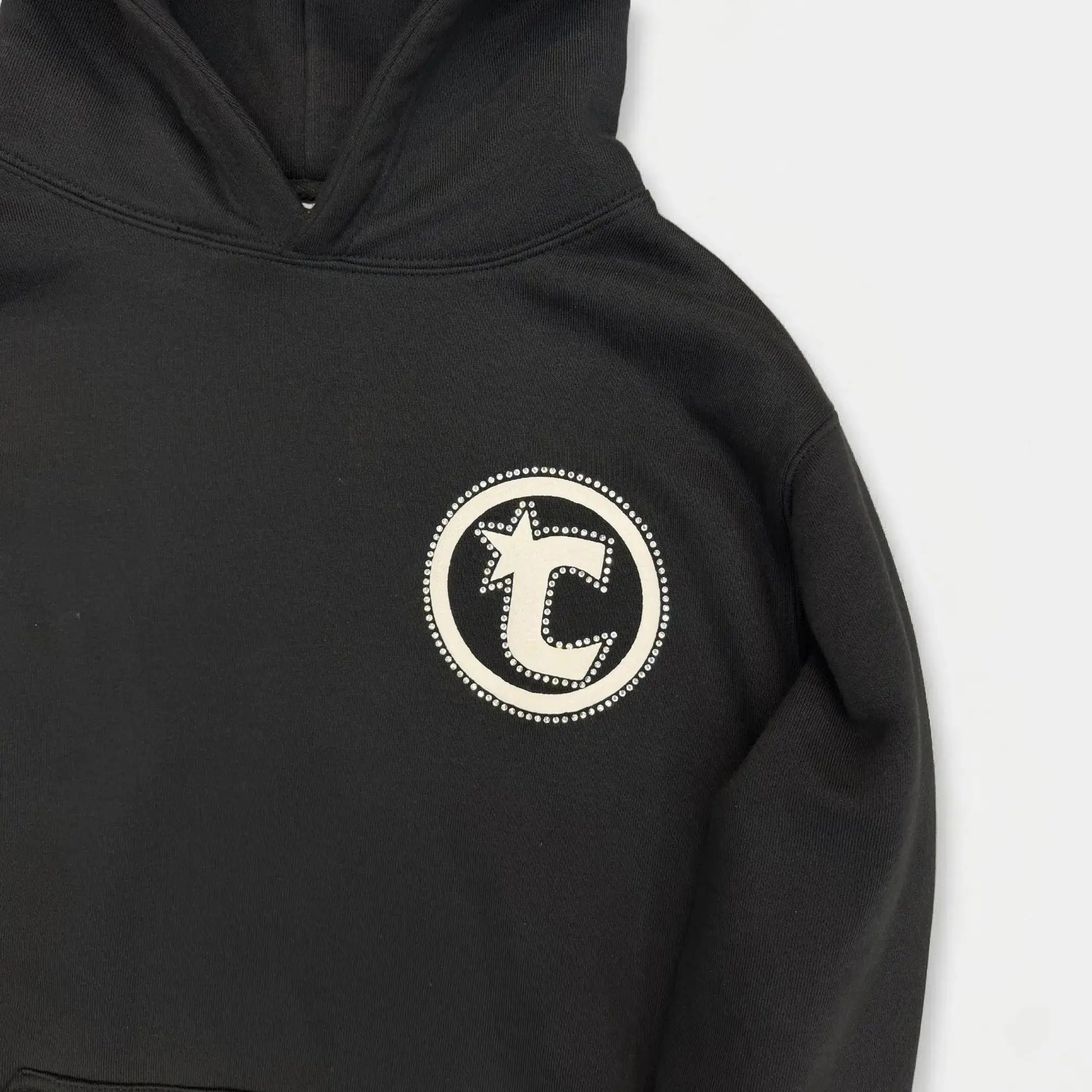 Extreme close-up of the CELESTIC Black Star Hoodie. Highlighting the prominent circular white 'C' star logo embroidery on the left chest with its intricate dotted border detail, set against the premium heavyweight black cotton fleece fabric texture.
