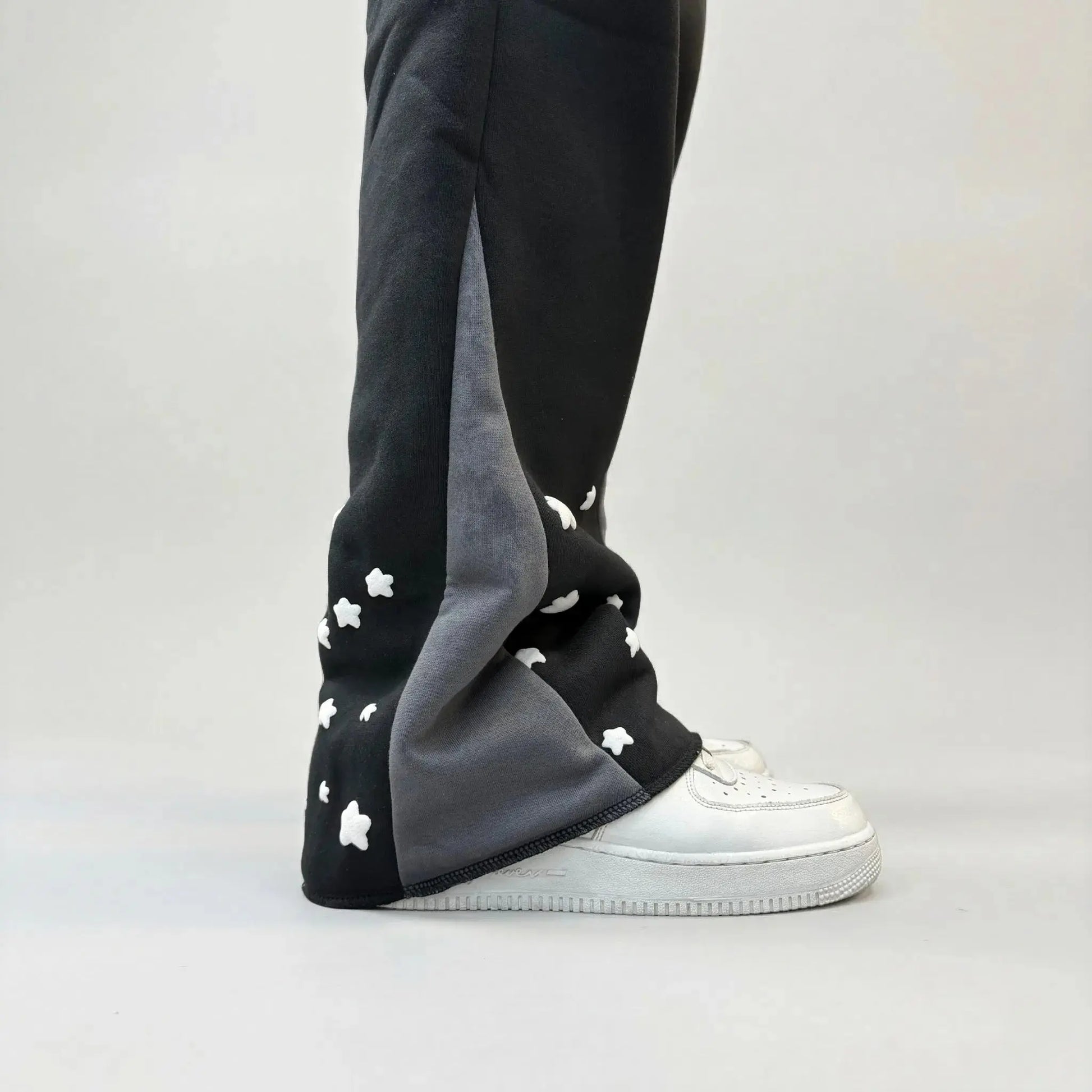 Detailed close-up of the lower leg on the CELESTIC Black Star sweatpants. Highlighting the premium heavyweight black cotton fleece, the contrasting grey side panel, and the scattered white embroidered star details as the leg rests on white sneakers.