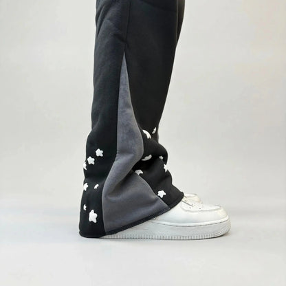 Detailed close-up of the lower leg on the CELESTIC Black Star sweatpants. Highlighting the premium heavyweight black cotton fleece, the contrasting grey side panel, and the scattered white embroidered star details as the leg rests on white sneakers.