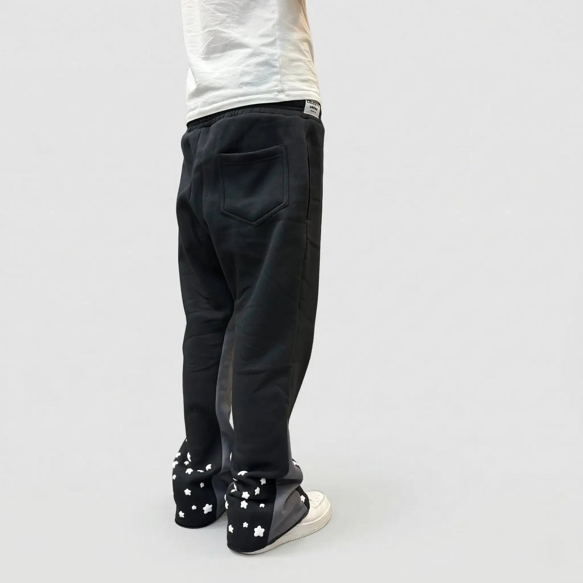 Back view of a model wearing the CELESTIC Black Star sweatpants. These premium heavyweight black cotton fleece pants feature contrasting grey side panels and multiple white embroidered stars scattered around the back of the leg openings. Shown in a relaxed straight-leg fit with white sneakers.