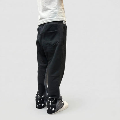 Back view of a model wearing the CELESTIC Black Star sweatpants. These premium heavyweight black cotton fleece pants feature contrasting grey side panels and multiple white embroidered stars scattered around the back of the leg openings. Shown in a relaxed straight-leg fit with white sneakers.