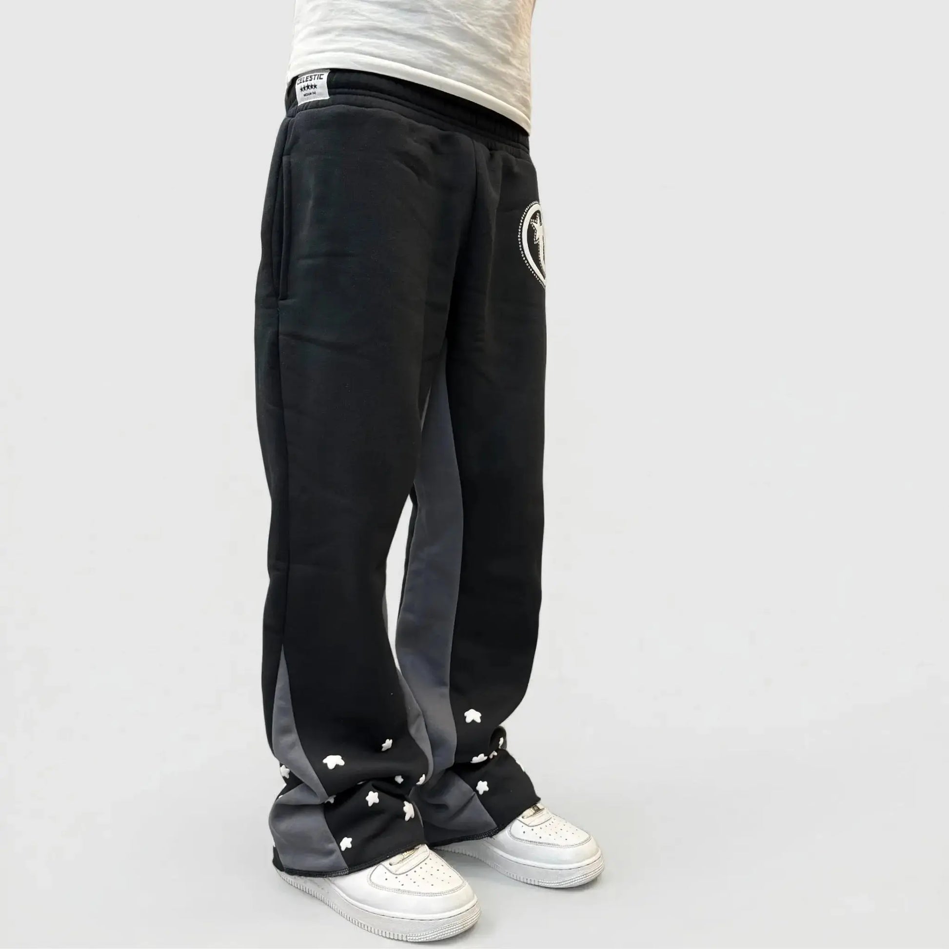 A detailed close-up and back-angle view of a model wearing the CELESTIC Black Star sweatpants. The black premium heavyweight cotton fleece pants feature large contrasting grey side panels and are adorned with multiple scattered white embroidered star details on the lower legs. Shown paired with white sneakers in a minimal studio setting.