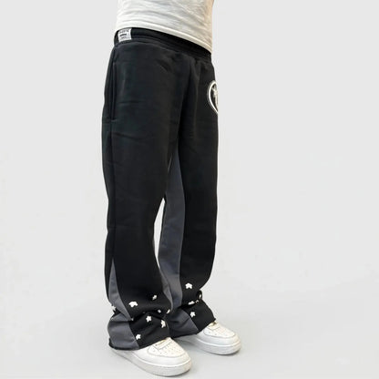 A detailed close-up and back-angle view of a model wearing the CELESTIC Black Star sweatpants. The black premium heavyweight cotton fleece pants feature large contrasting grey side panels and are adorned with multiple scattered white embroidered star details on the lower legs. Shown paired with white sneakers in a minimal studio setting.