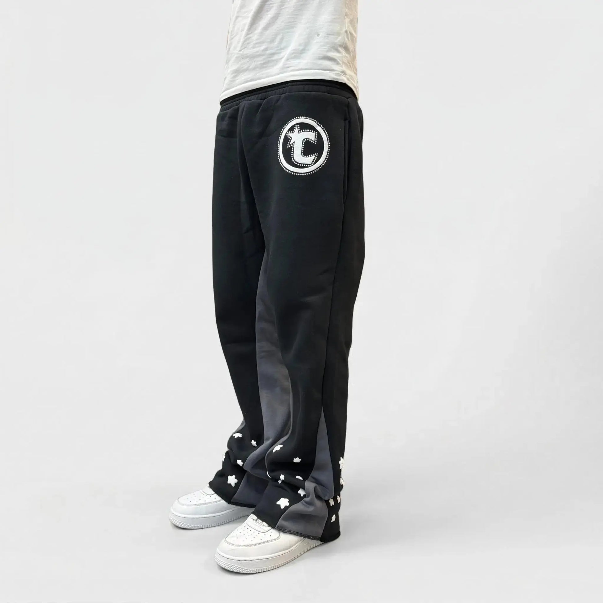 Side view of a model wearing the CELESTIC Black Star sweatpants. These premium heavyweight black cotton fleece pants feature unique grey side panels, a large white circular 'C' star logo on the thigh, and multiple white embroidered stars around the leg openings. Shown with white sneakers on a light background.