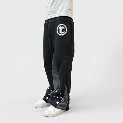 Side view of a model wearing the CELESTIC Black Star sweatpants. These premium heavyweight black cotton fleece pants feature unique grey side panels, a large white circular 'C' star logo on the thigh, and multiple white embroidered stars around the leg openings. Shown with white sneakers on a light background.