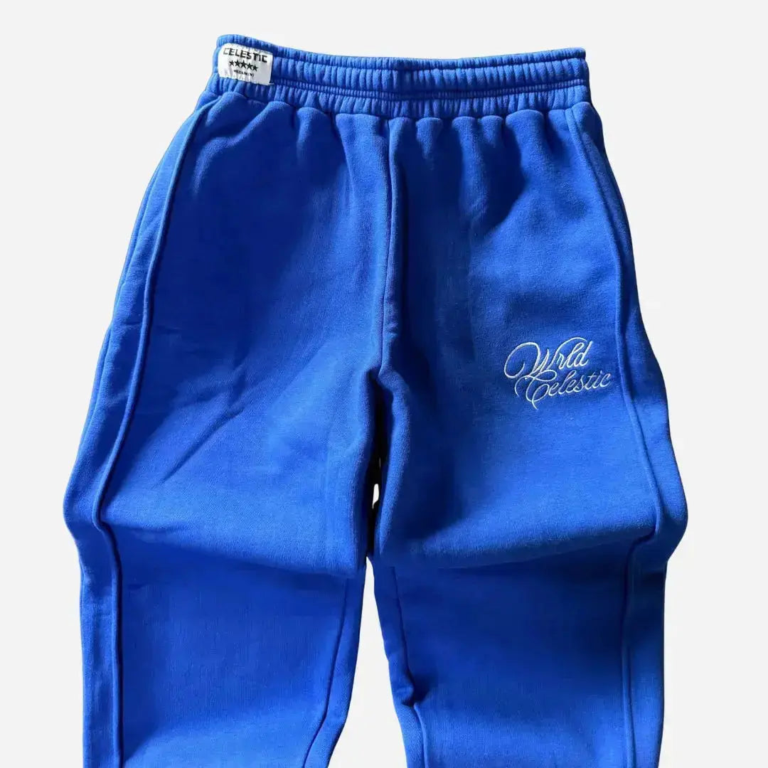 Detailed close-up of the upper front of the CELESTIC Blue Basic sweatpants. Highlighting the elasticated ribbed waistband with a woven brand tag, prominent side seams, and the elegant white script logo embroidery on the left thigh against the premium heavyweight blue fleece.