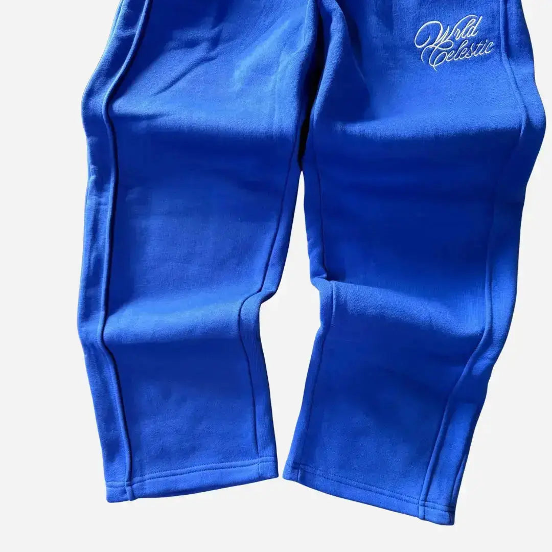 Detailed close-up of the lower legs and hems of the CELESTIC Blue Basic sweatpants. Highlighting the relaxed straight fit with an open hem, prominent side panel seams, and the elegant white script logo embroidery on the thigh against the premium heavyweight blue fleece.