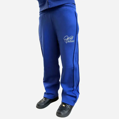 Blue oversized tracksuit pants angle view on model. Premium heavyweight cotton fleece sweatpants from the CELESTIC Blue Basic set, highlighting the white script logo embroidery on the left thigh, prominent side seam detailing, and the relaxed fit stacking perfectly over black sneakers.