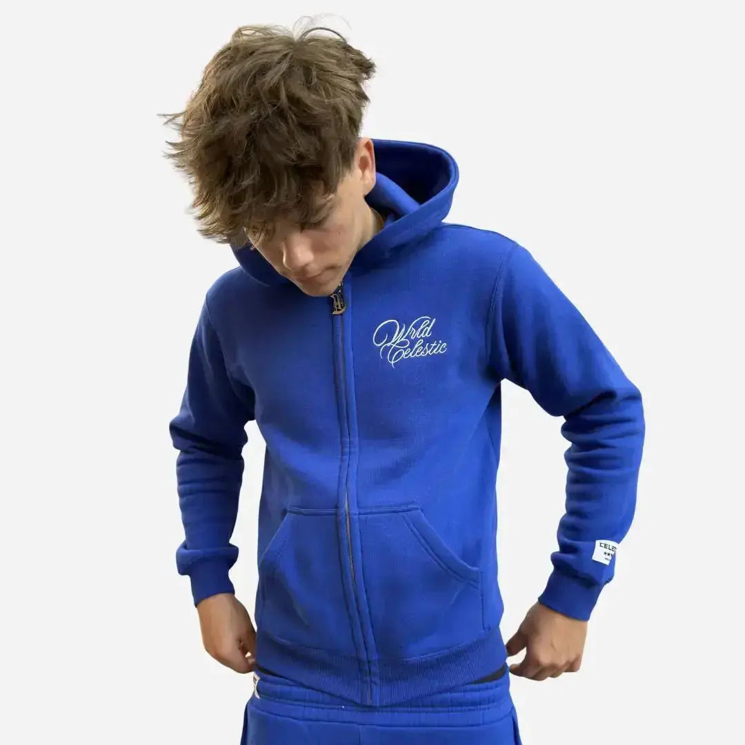 Front view of a model wearing the CELESTIC Blue Basic Tracksuit. Highlighting the premium heavyweight blue fleece oversized zip-up hoodie, featuring the white script logo chest embroidery, custom metal zipper pull, and the woven brand patch on the sleeve.