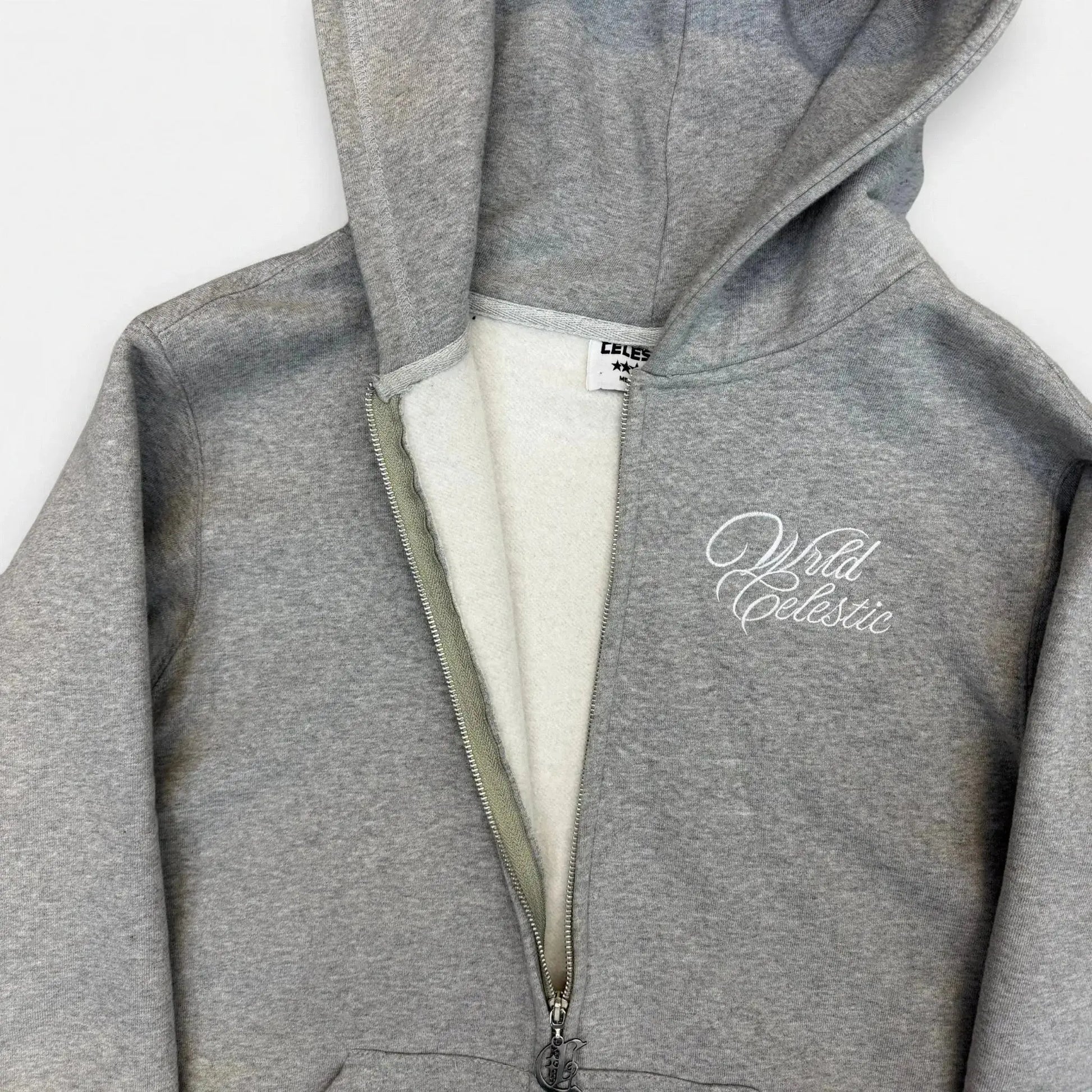 Detailed close-up of the CELESTIC Grey Basic zip-up hoodie, partially unzipped to show the soft inner fleece lining. Highlighting the premium heavyweight grey cotton fabric, the custom metal 'C' zipper pull, and the elegant white script logo embroidery on the chest.