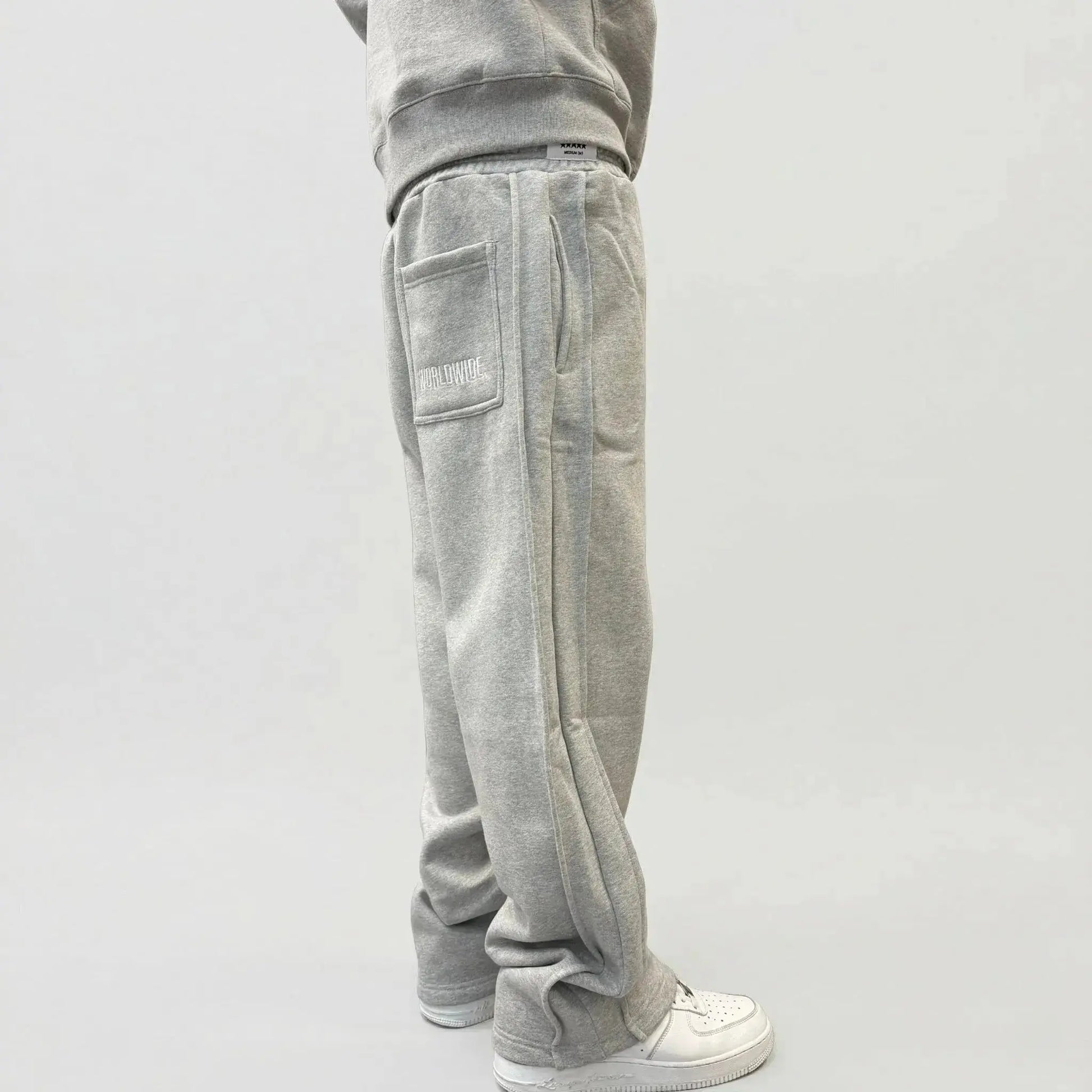 Back angle view of a model wearing the CELESTIC Grey Basic sweatpants. These premium heavyweight grey cotton fleece pants feature a relaxed straight-leg silhouette, white 'WORLDWIDE' embroidery on the right back pocket, and prominent side panel seams. Shown with white sneakers on a clean light background.