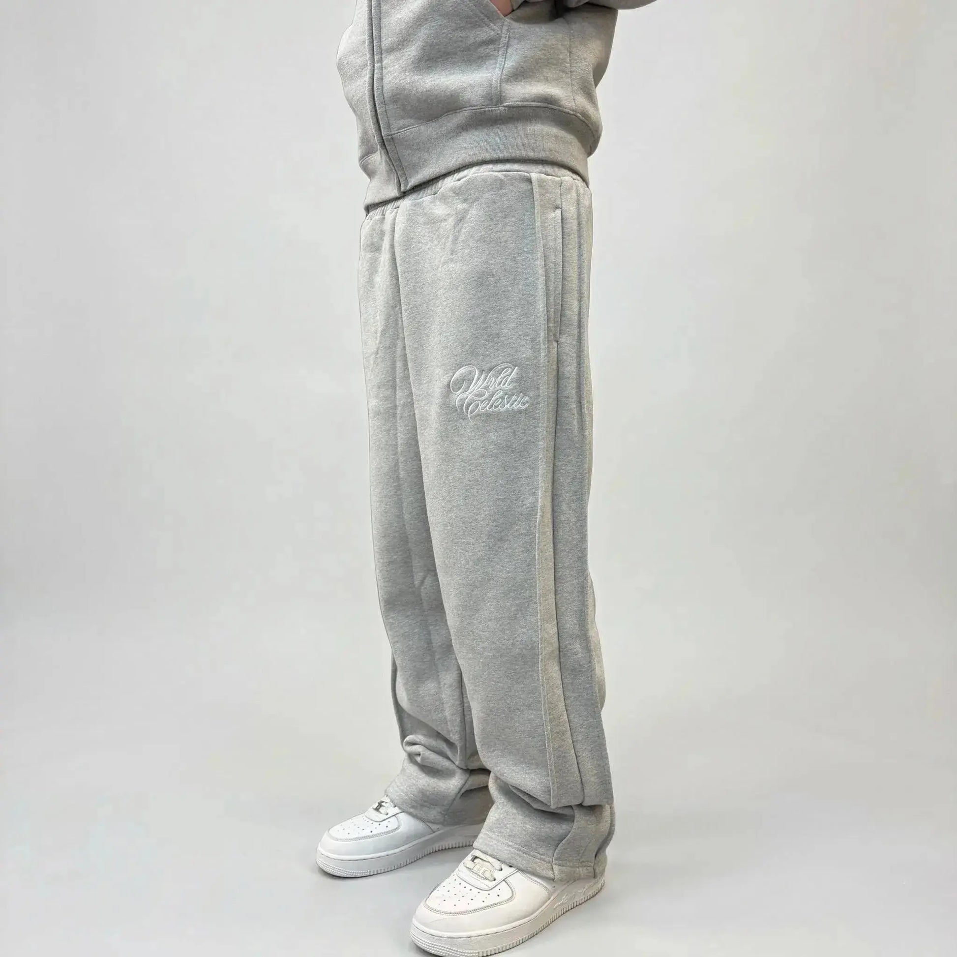 Side view of a model wearing the CELESTIC Grey Basic sweatpants. These premium heavyweight grey cotton fleece pants feature a relaxed straight-leg fit, prominent side panel seams, and elegant white script logo embroidery on the left thigh. Shown paired with white sneakers.
