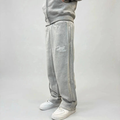 Side view of a model wearing the CELESTIC Grey Basic sweatpants. These premium heavyweight grey cotton fleece pants feature a relaxed straight-leg fit, prominent side panel seams, and elegant white script logo embroidery on the left thigh. Shown paired with white sneakers.