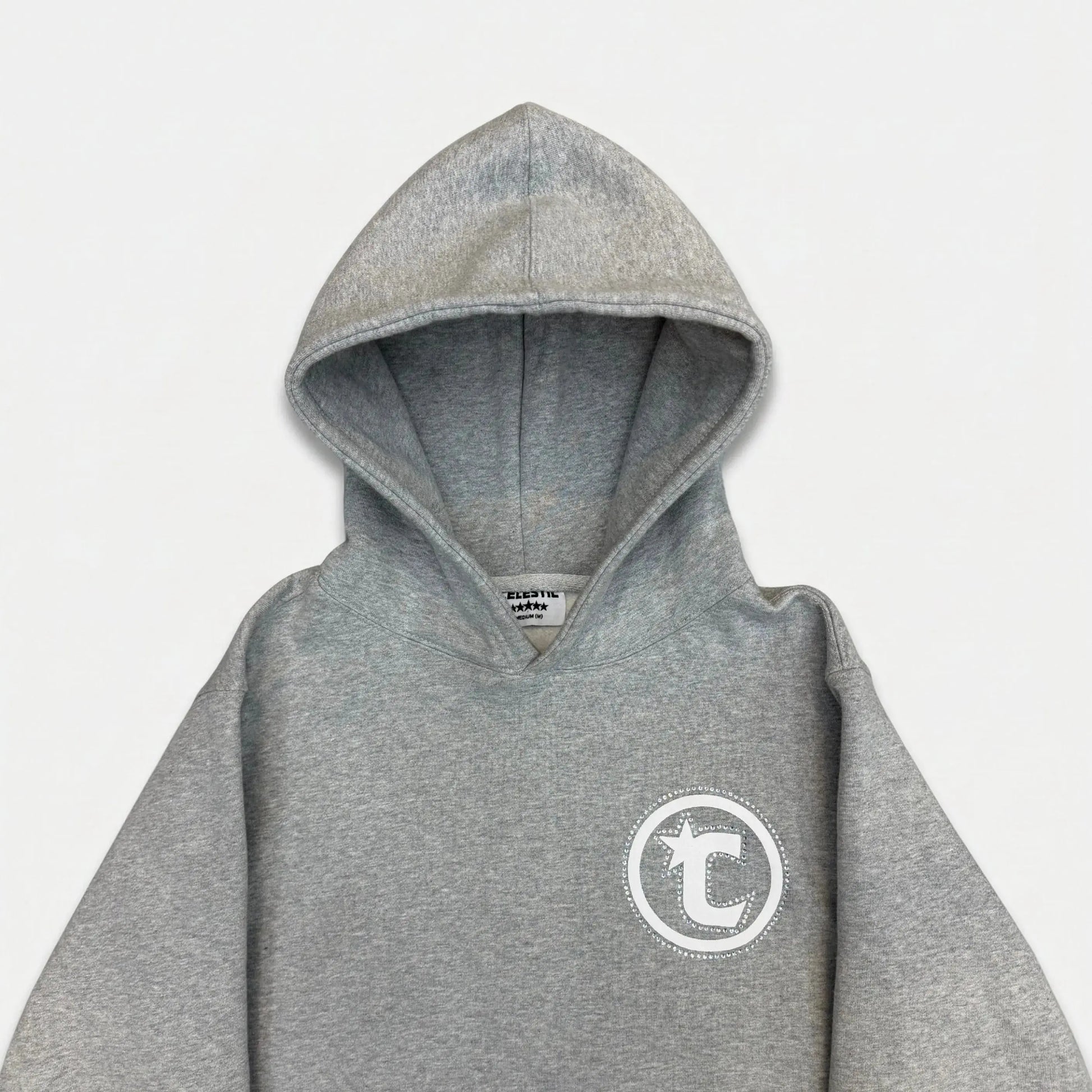 Front view flat-lay photograph of the CELESTIC Grey Star Hoodie on a white background. This premium heather grey cotton fleece pullover hoodie features light grey side panels and a spacious kangaroo pocket. A large white circular 'C' star logo is screen-printed on the left chest. The sleeves and lower body show white embroidered star details. A white woven brand label is visible on the inner neck.