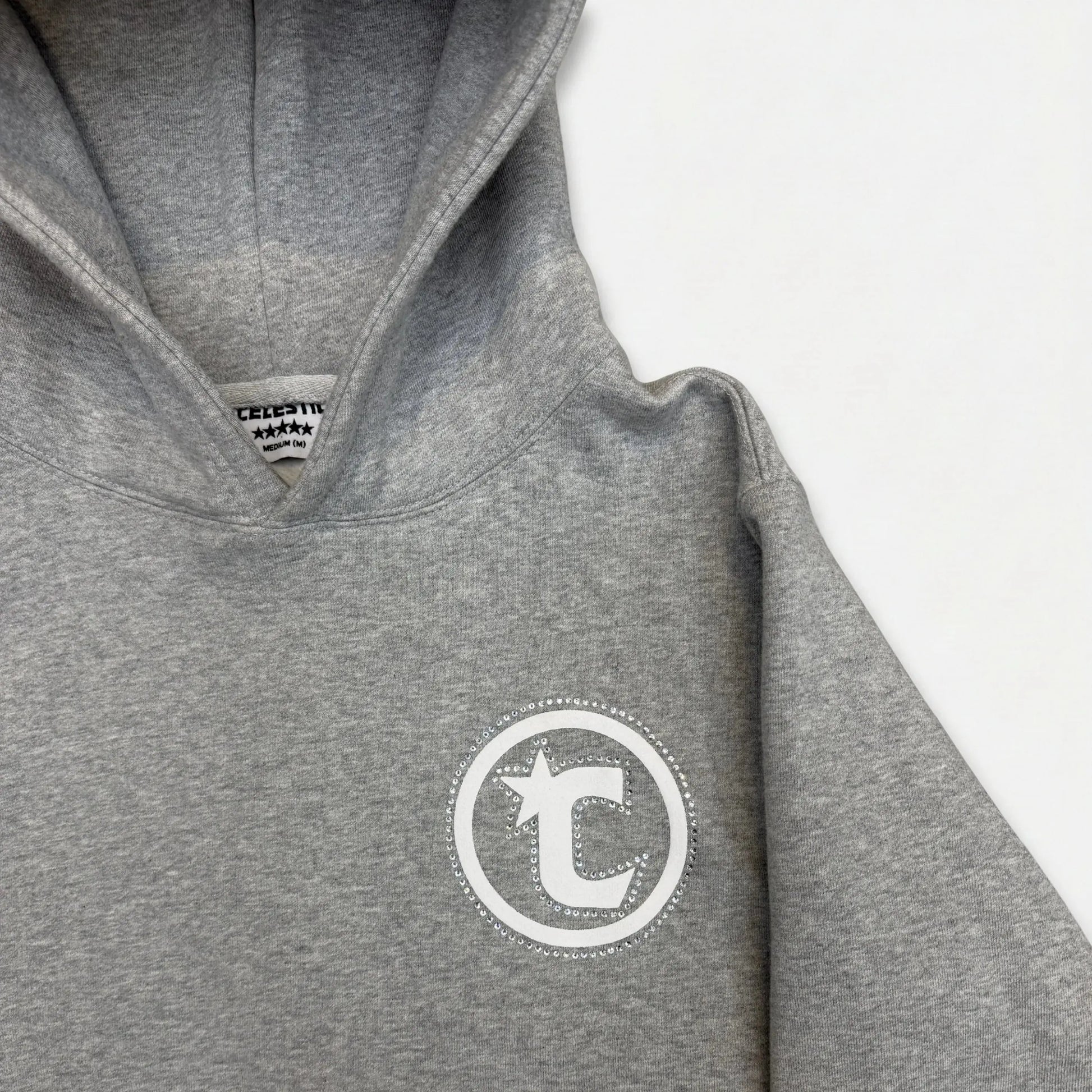 A detailed close-up photograph of the upper section of the CELESTIC Grey Star Hoodie. The image highlights the structured double-lined hood and the premium heather grey cotton fleece fabric. On the left chest, a white circular 'C' star logo is featured, elegantly framed by a border of sparkling rhinestones. The internal CELESTIC brand neck label is partially visible.