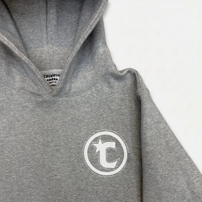 A detailed close-up photograph of the upper section of the CELESTIC Grey Star Hoodie. The image highlights the structured double-lined hood and the premium heather grey cotton fleece fabric. On the left chest, a white circular 'C' star logo is featured, elegantly framed by a border of sparkling rhinestones. The internal CELESTIC brand neck label is partially visible.