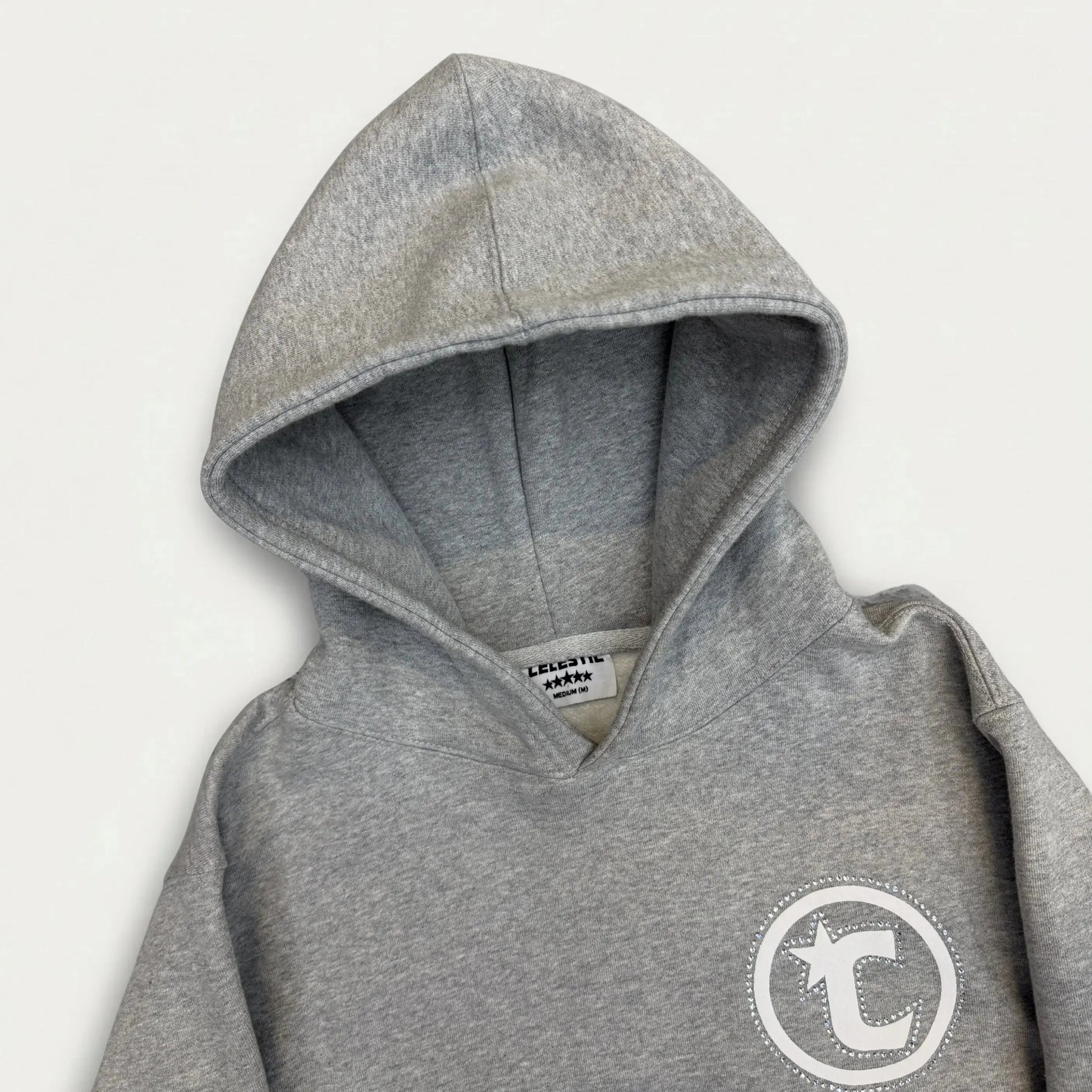 A detailed close-up photograph of the CELESTIC Grey Star Hoodie on a white background. The image focuses on the structured double-lined hood and the chest area, showcasing the premium heather grey cotton fleece fabric. On the left chest, a white circular 'C' star logo is featured, elegantly framed by a border of shimmering rhinestones. The internal CELESTIC brand neck label is visible at the base of the hood.