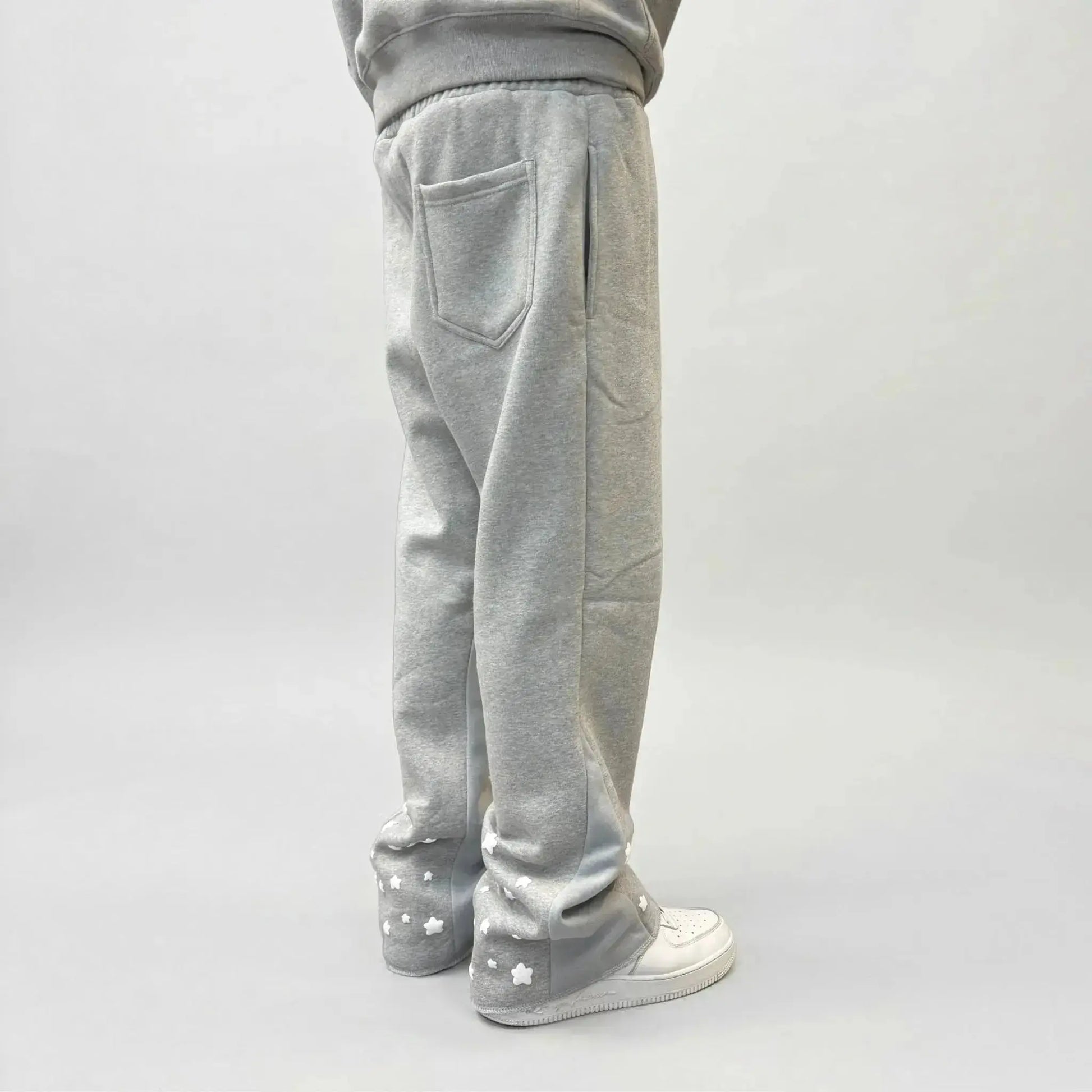 On-body rear view of the CELESTIC Grey Star Sweatpants, styled with white sneakers. These premium heather grey cotton fleece sweatpants feature a relaxed fit, a single back pocket on the right side, and light grey side panels. The lower back of both legs is detailed with multiple scattered white embroidered stars, showcasing the unique streetwear aesthetic of the Star collection.