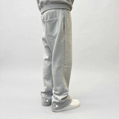 On-body rear view of the CELESTIC Grey Star Sweatpants, styled with white sneakers. These premium heather grey cotton fleece sweatpants feature a relaxed fit, a single back pocket on the right side, and light grey side panels. The lower back of both legs is detailed with multiple scattered white embroidered stars, showcasing the unique streetwear aesthetic of the Star collection.