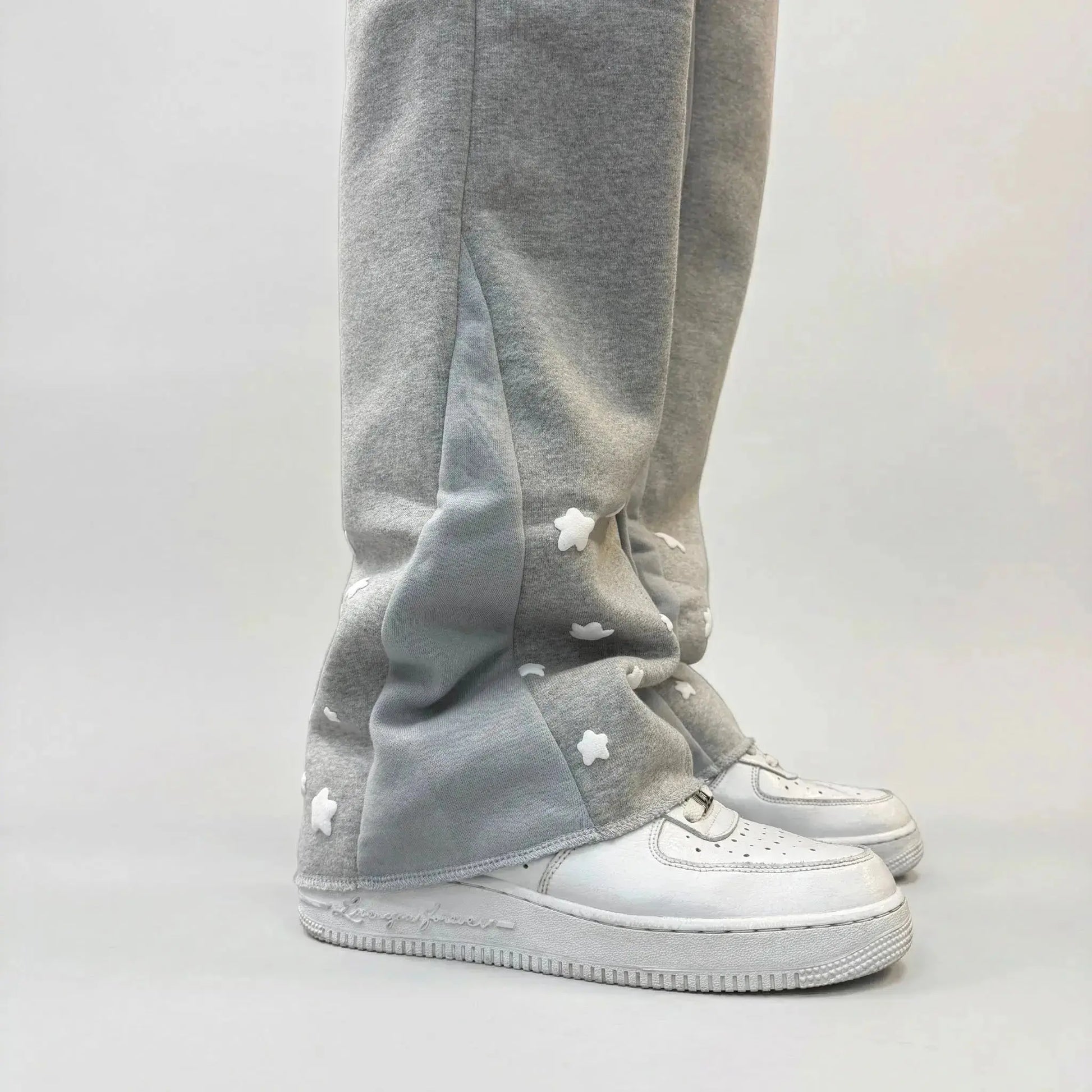 A detailed close-up photograph of the lower leg of the CELESTIC Grey Star Sweatpants, styled with a white leather sneaker. The shot highlights the premium heather grey cotton fleece fabric and the light grey side panel. Multiple white embroidered stars are scattered across the hem area, showcasing the intricate craftsmanship and unique streetwear design of the Star collection.