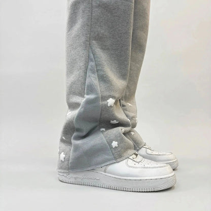 A detailed close-up photograph of the lower leg of the CELESTIC Grey Star Sweatpants, styled with a white leather sneaker. The shot highlights the premium heather grey cotton fleece fabric and the light grey side panel. Multiple white embroidered stars are scattered across the hem area, showcasing the intricate craftsmanship and unique streetwear design of the Star collection.
