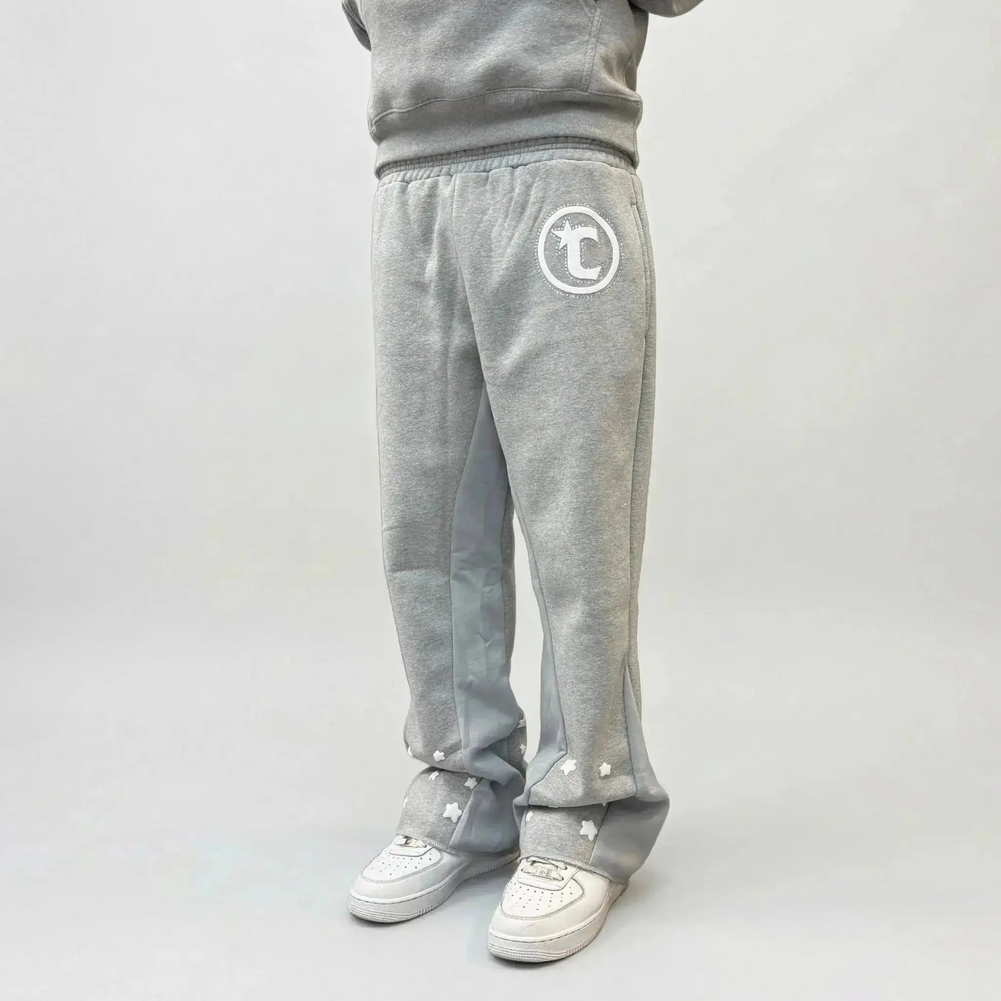 On-body side view of a person wearing the CELESTIC Grey Star Sweatpants, styled with white sneakers. The premium heather grey cotton fleece sweatpants feature a relaxed fit, light grey side panels, and a white circular 'C' star logo on the left thigh. Multiple white embroidered stars are scattered across the lower leg and hem area, highlighting the unique design details of the Star collection.