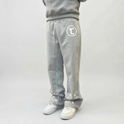 On-body side view of a person wearing the CELESTIC Grey Star Sweatpants, styled with white sneakers. The premium heather grey cotton fleece sweatpants feature a relaxed fit, light grey side panels, and a white circular 'C' star logo on the left thigh. Multiple white embroidered stars are scattered across the lower leg and hem area, highlighting the unique design details of the Star collection.