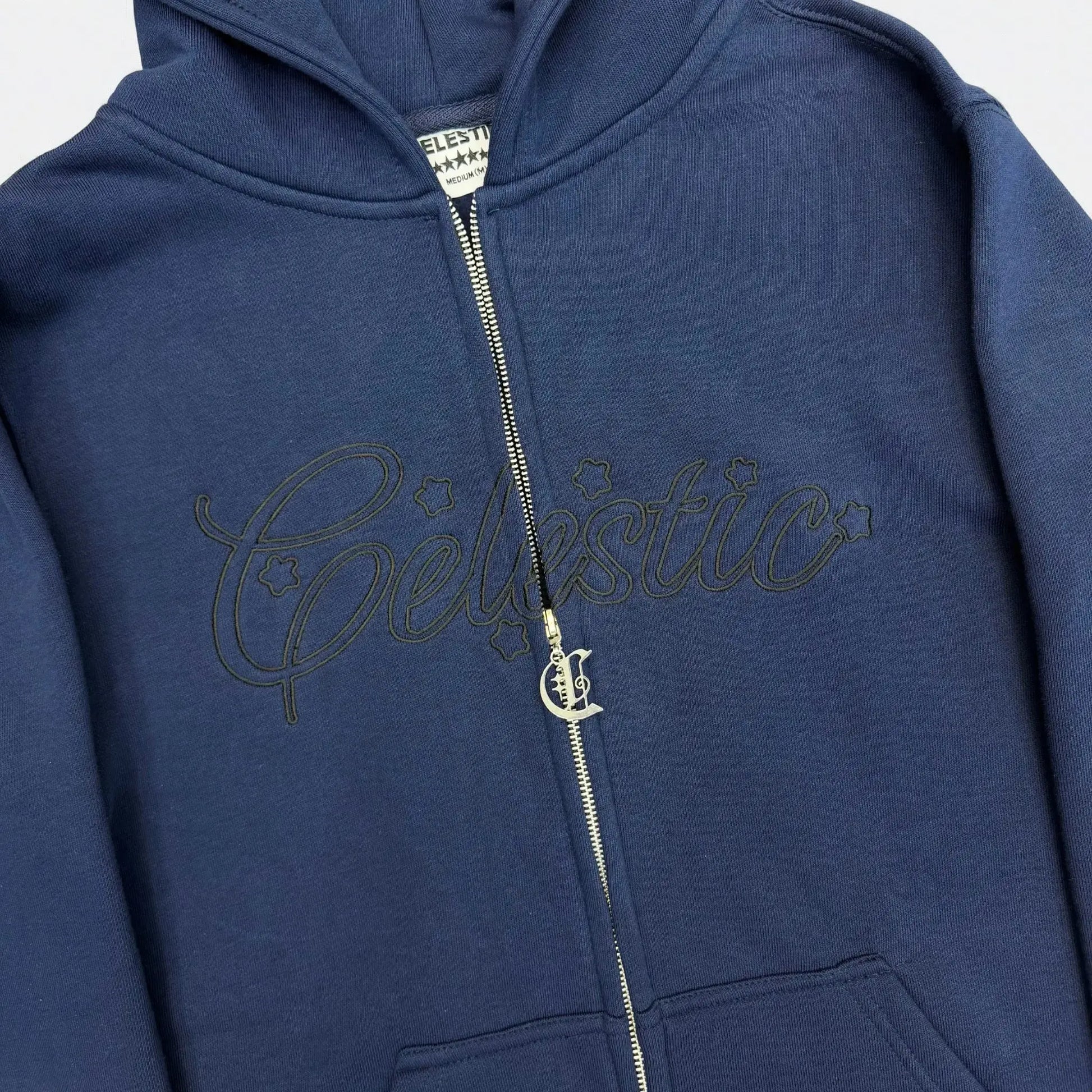 Detailed close-up of the CELESTIC Navy Blue Outline zip-up hoodie. Featuring the large tonal 'Celestic' outline embroidery with star details, the custom silver metal 'C' zipper pull, and the premium heavyweight navy blue cotton fleece fabric.