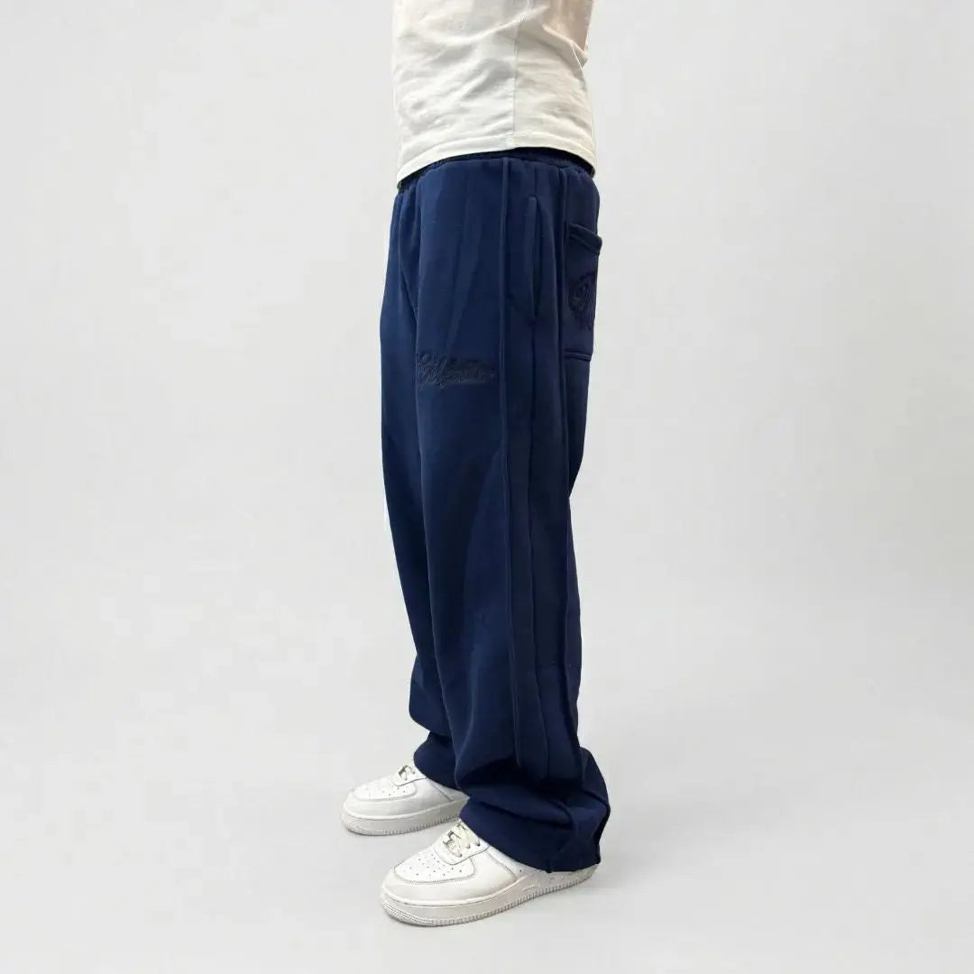 Side view of a model wearing the CELESTIC Navy Blue Outline sweatpants. These premium heavyweight cotton fleece pants feature a relaxed straight-leg fit, prominent side panel seams, and tonal 'Celestic' outline embroidery on the left thigh and back pocket. Shown paired with white sneakers.