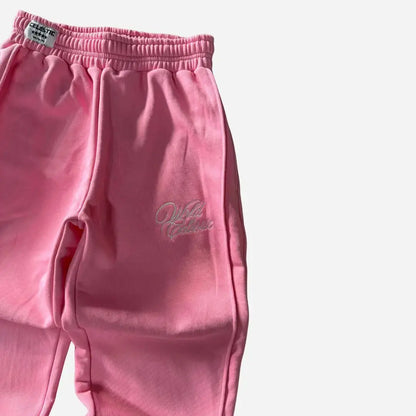 Angled close-up view of the CELESTIC Pink Basic sweatpants focusing on the left side. Highlighting the elegant white script logo embroidery on the thigh, prominent side seam detailing, and the elasticated ribbed waistband with a woven brand tag against the premium heavyweight pink fleece.