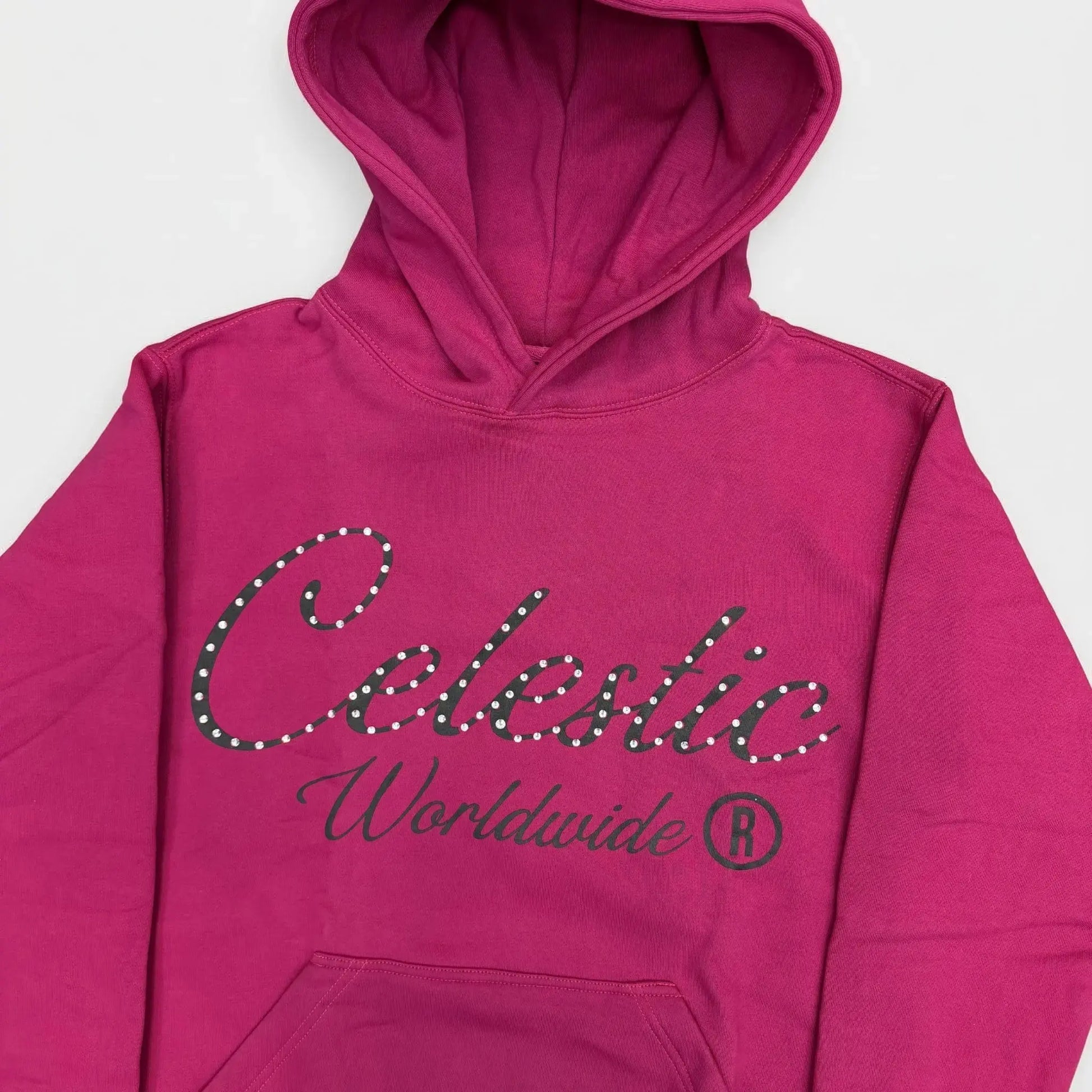 Detailed close-up photograph of the CELESTIC Pink Gemstone Hoodie. The focus is on the chest, showcasing the vibrant pink premium cotton fleece and the large black screen-printed 'Celestic' script logo, heavily detailed with sparkling rhinestones. The printed text 'Worldwide ®' is clearly visible below the script. The base of the structured hood and the top of the kangaroo pocket frame the logo.