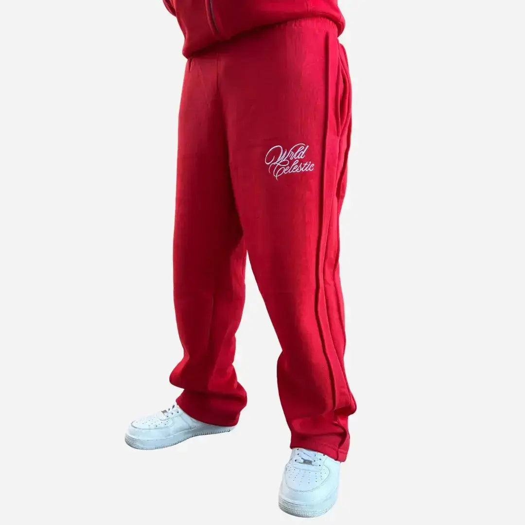 Red oversized tracksuit pants angle view on model. Premium heavyweight cotton fleece sweatpants from the CELESTIC Red Basic set, highlighting the white script logo embroidery on the left thigh, prominent side seam detailing, and the relaxed fit stacking perfectly over white sneakers.