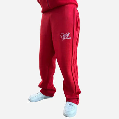 Red oversized tracksuit pants angle view on model. Premium heavyweight cotton fleece sweatpants from the CELESTIC Red Basic set, highlighting the white script logo embroidery on the left thigh, prominent side seam detailing, and the relaxed fit stacking perfectly over white sneakers.